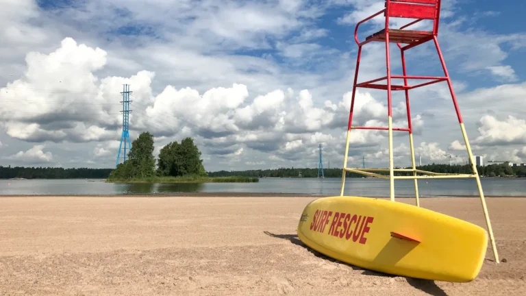 Glābēju tornis un dzeltena glābšanas laiva smilšainā pludmalē zem mākoņainām debesīm.