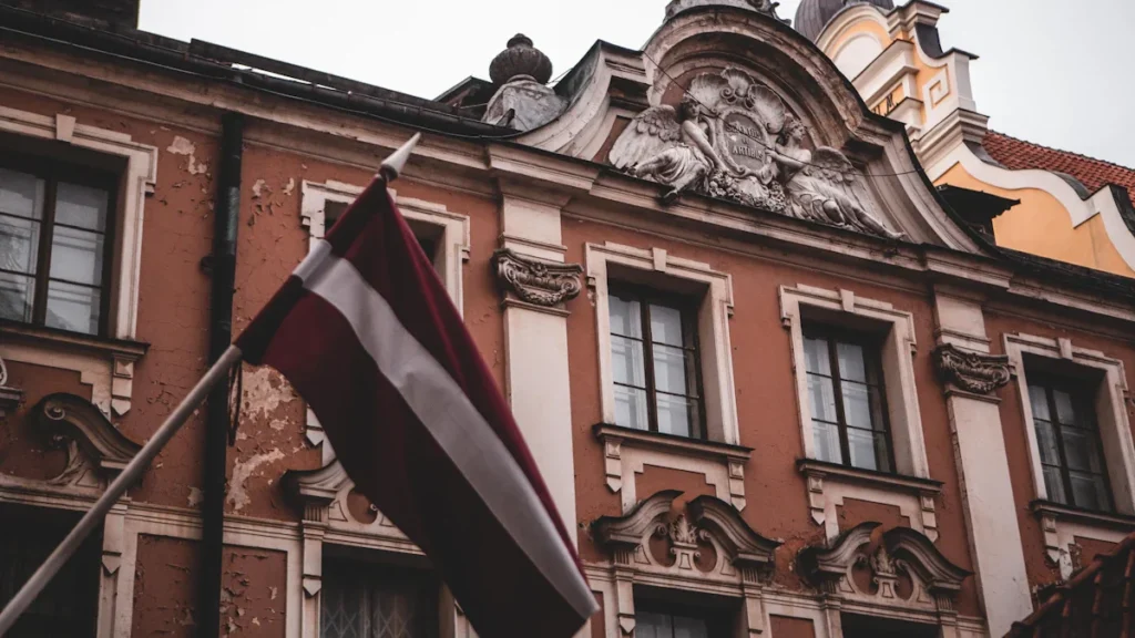 Latvijas valsts karogs plīvo pie vēsturiskas ēkas Rīgas centrā, simbolizējot nacionālo piederību un vēlēšanas.
