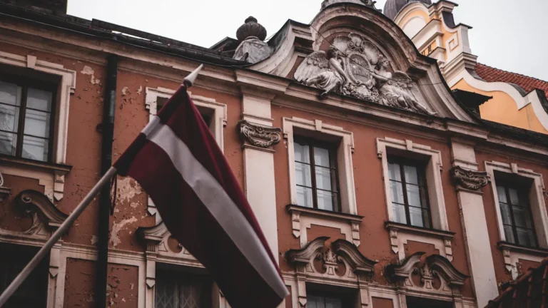 Latvijas valsts karogs plīvo pie vēsturiskas ēkas Rīgas centrā, simbolizējot nacionālo piederību un vēlēšanas.