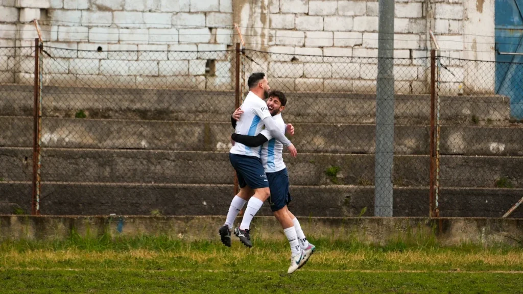 Divi futbola spēlētāji gaišos sporta tērpos emocionāli svin gūtos vārtus uz laukuma.