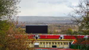 Futbola stadions ar sarkanām sēdvietām un lielu ekrānu vēsturiskā rezultāta paziņošanai.