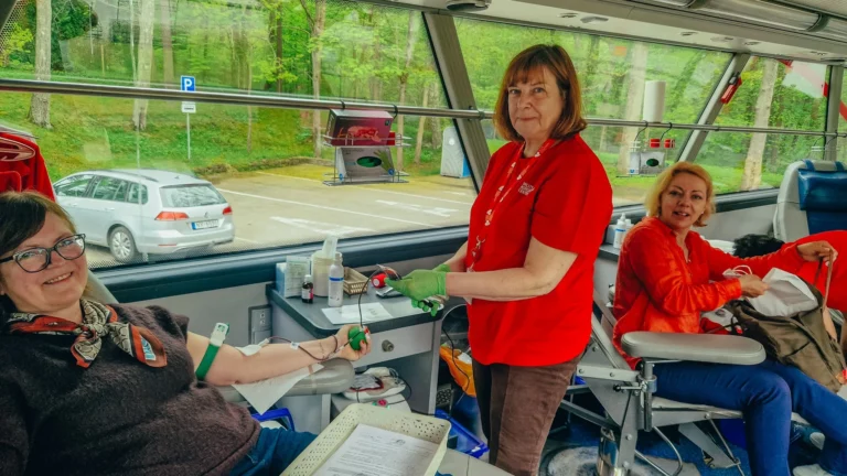 Sieviete sēž krēslā un ziedo asinis mobilajā autobusā, kamēr medicīnas darbiniece sarkanā formā viņai palīdz.