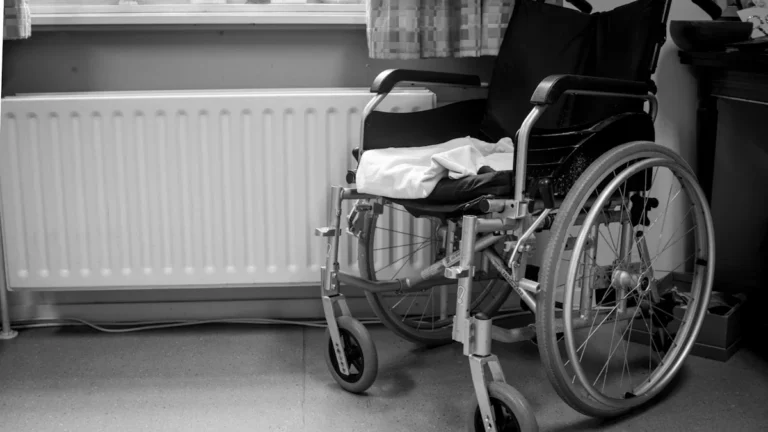Invalīdu ratiņkrēsls novietots telpā blakus radiatoram, simbolizējot sociālo aprūpi un ikdienas atbalstu.