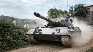 Kaujas tanks pārvietojas pa mācību lauku, radot putekļu mākoni un troksni meža ielokā.