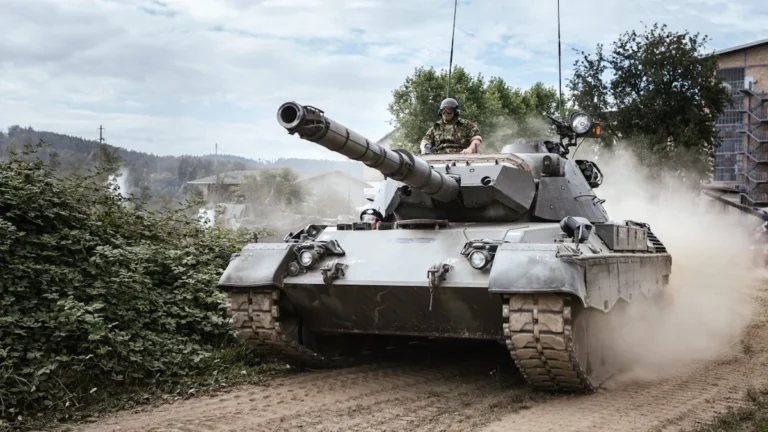 Kaujas tanks pārvietojas pa mācību lauku, radot putekļu mākoni un troksni meža ielokā.