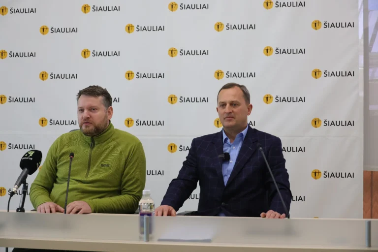 Divi vīrieši sēž pie galda uz balta fona ar Šauļu pilsētas logotipiem un mikrofoniem.