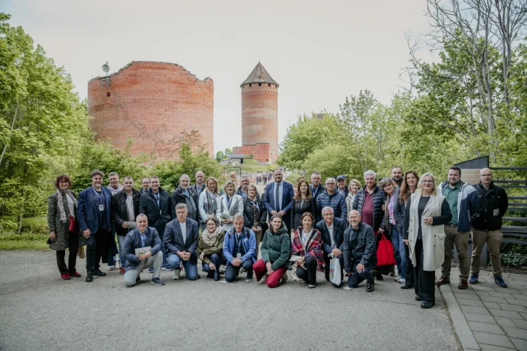 Cilvēku grupa pozē fotoattēlam pie Turaidas pils sarkanajiem ķieģeļu torņiem un zaļiem kokiem.