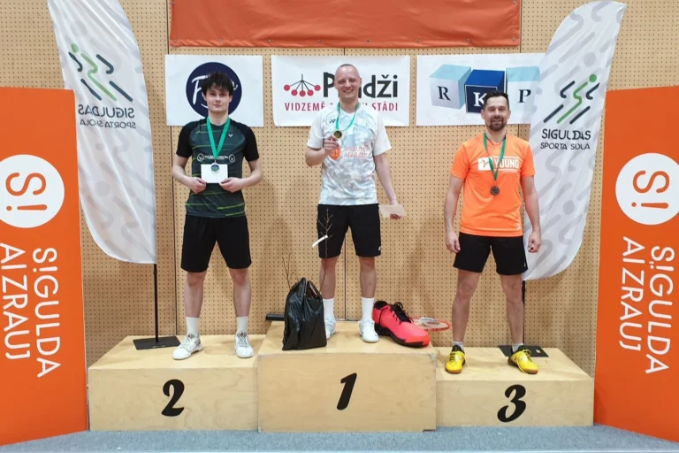 Trīs vīrieši ar medaļām kaklā stāv uz goda pjedestāla sporta hallē pie sponsoru baneriem.
