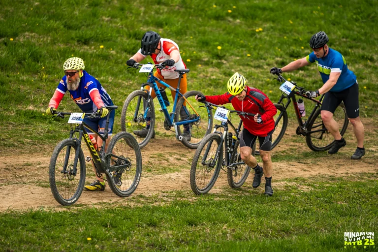 Četri kalnu riteņbraucēji stumj savus velosipēdus augšup pa stāvu, zāļainu nogāzi sacensību laikā.
