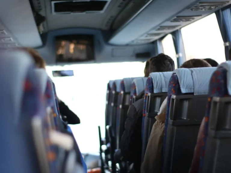 Skats uz autobusa salonu no aizmugures, kur sēž pasažieri un priekšā redzams neliels ekrāns.