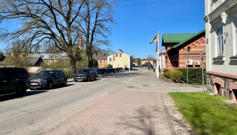 Ielas skats ar automašīnām, kokiem un vēsturiskām ēkām saulainā dienā zem skaidrām debesīm.