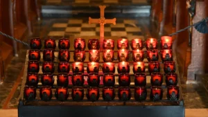 Degošas lūgšanu svecītes sarkanos trauciņos pie krusta uz tumša baznīcas interjera fona.