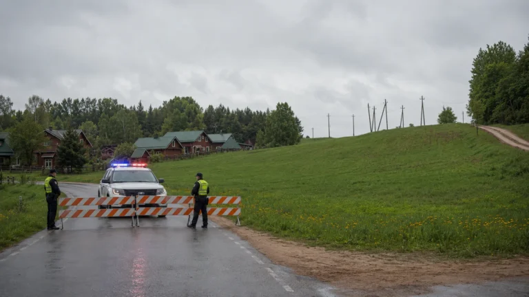 Divi policisti un policijas automašīna ar barjerām bloķē lauku ceļu pie dzīvojamām mājām.