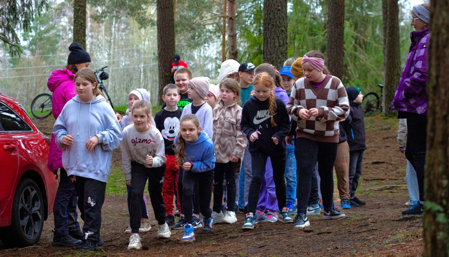 Bērnu grupa krāsainā sporta apģērbā stāv uz meža takas, gatavojoties startam skriešanas sacensībās.