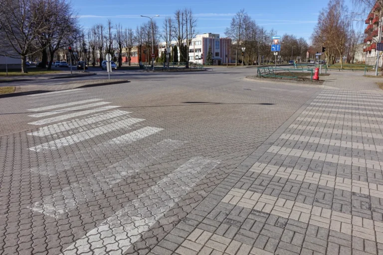 Bruģēts krustojums ar gājēju pāreju, kailiem kokiem un ēkām zem skaidrām, zilām debesīm.