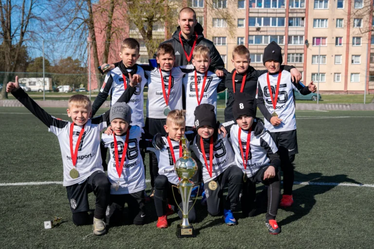 Jauniešu futbola komanda baltos kreklos ar medaļām un lielu zelta kausu pozē uz mākslīgā zālāja.