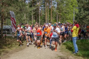 Skrējēju grupa ar starta numuriem enerģiski uzsāk skriešanas sacensības pa meža taku starp priedēm.
