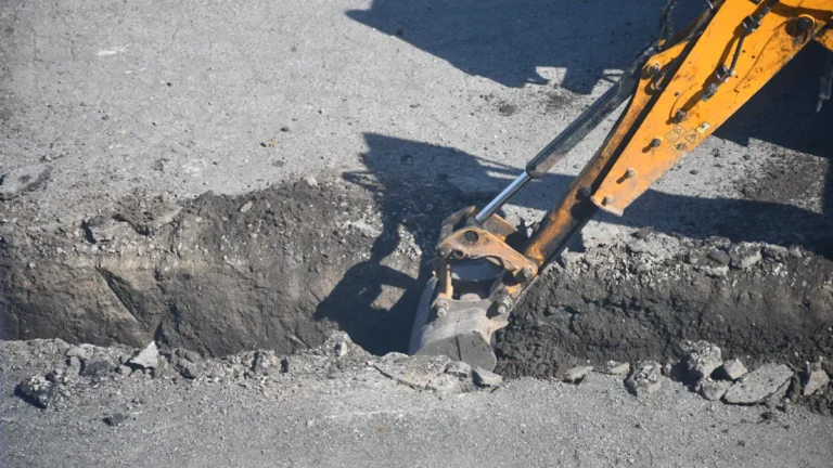 Ekskavators rok tranšeju ceļa segumā jaunā elektropārvades kabeļa izbūves darbu laikā.