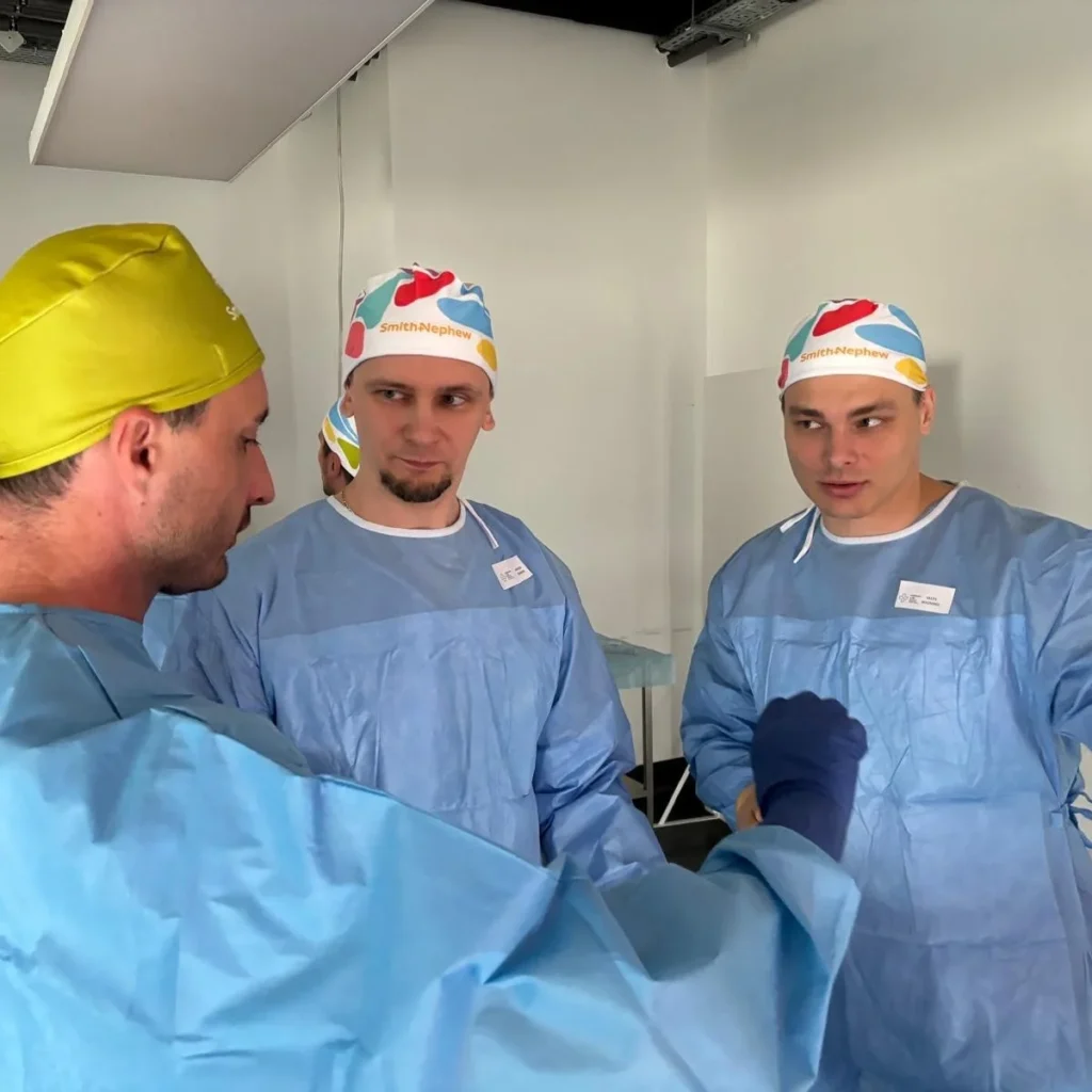 Trīs vīrieši gaiši zilos medicīnas halātos un krāsainās cepurēs apspriežas telpā ar baltām sienām.