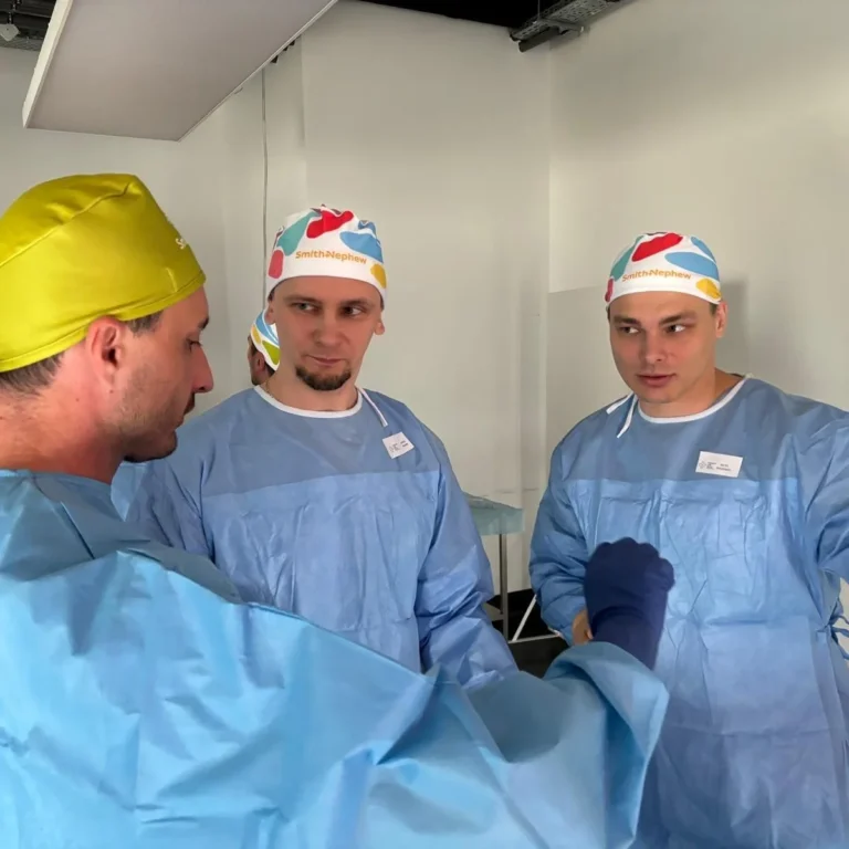 Trīs vīrieši gaiši zilos medicīnas halātos un krāsainās cepurēs apspriežas telpā ar baltām sienām.