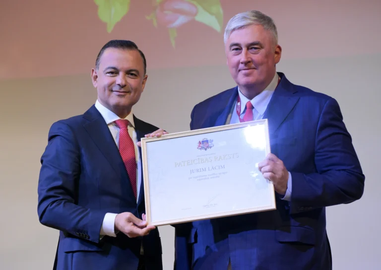 Divi vīrieši tumšos uzvalkos un sarkanās kaklasaitēs kopā tur lielu, baltu ierāmētu pateicības rakstu.