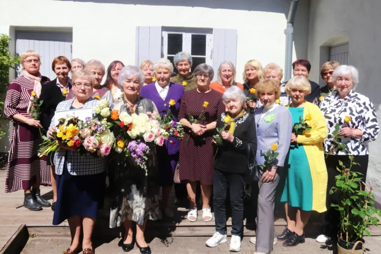 Grupas portrets ar sirmgalvju sievietēm, kuras smaidot stāv pie ēkas un tur rokās ziedus.