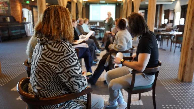 Sieviete rakstainā jakā un citi dalībnieki sēž konferenču zālē, klausoties lektora prezentāciju tālumā.