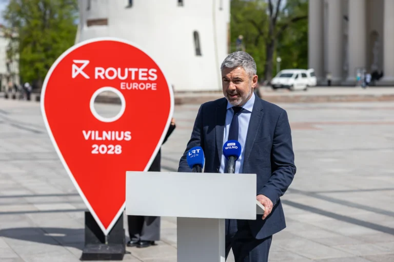 Vīrietis uzvalkā uzstājas pie pults, blakus redzama liela sarkana 'Routes Europe Vilnius 2028' norādes zīme.