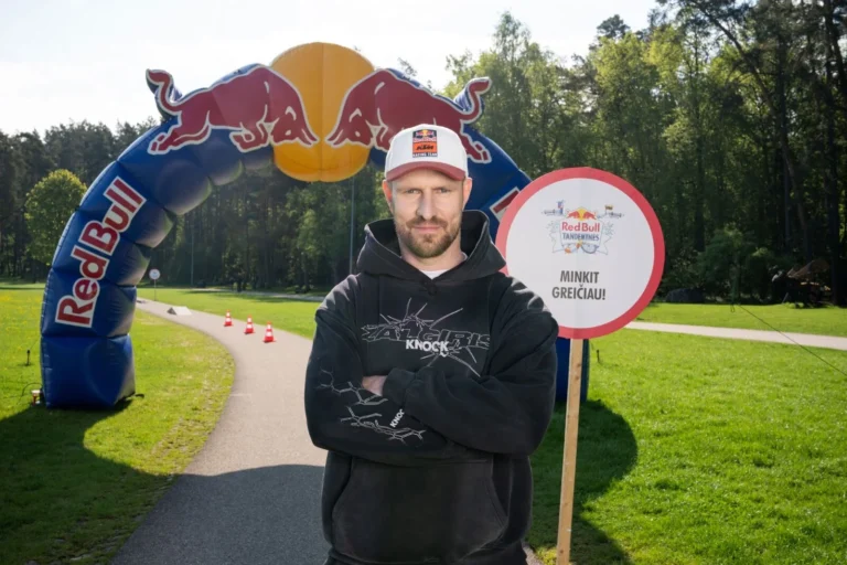 Vīrietis ar sakrustotām rokām stāv pie Red Bull arkas un pasākuma zīmes saulainā parkā.