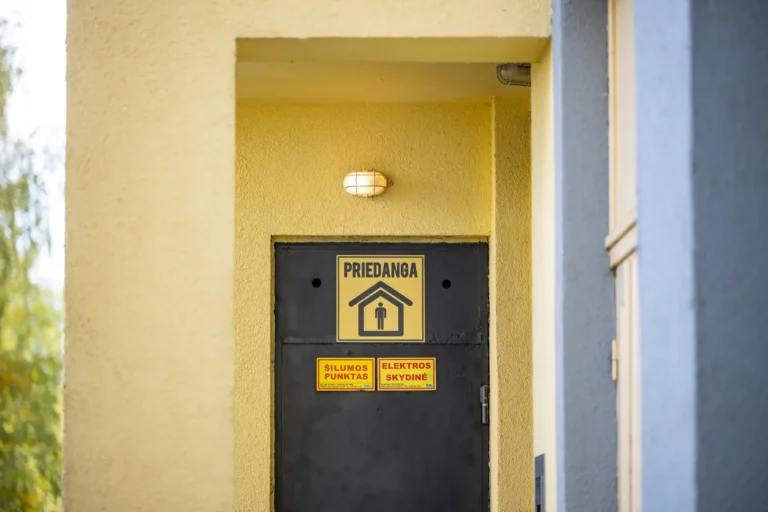 Melnas metāla durvis dzeltenā ēkas sienā ar zīmi, kas apzīmē patvēruma vietu vai patvertni.