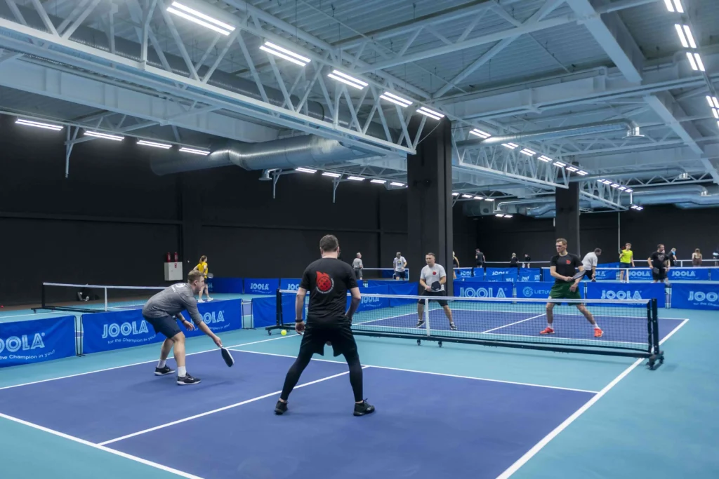 Vīrieši sporta zālē spēlē pikelbolu uz ziliem laukumiem ar JOOLA aprīkojumu un tīkliem.