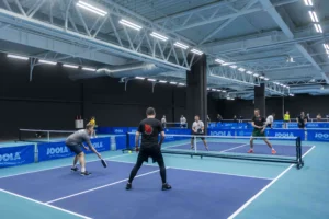 Vīrieši sporta zālē spēlē pikelbolu uz ziliem laukumiem ar JOOLA aprīkojumu un tīkliem.