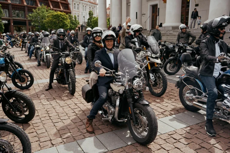 Eleganti tērpti motociklisti uz klasiskiem motocikliem pulcējas pilsētas laukumā uz bruģēta seguma.