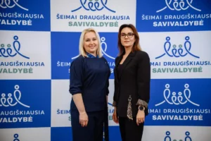 Divas smaidīgas sievietes lietišķā apģērbā pozē pie zili balta informatīvā stenda ar pasākuma logotipiem.