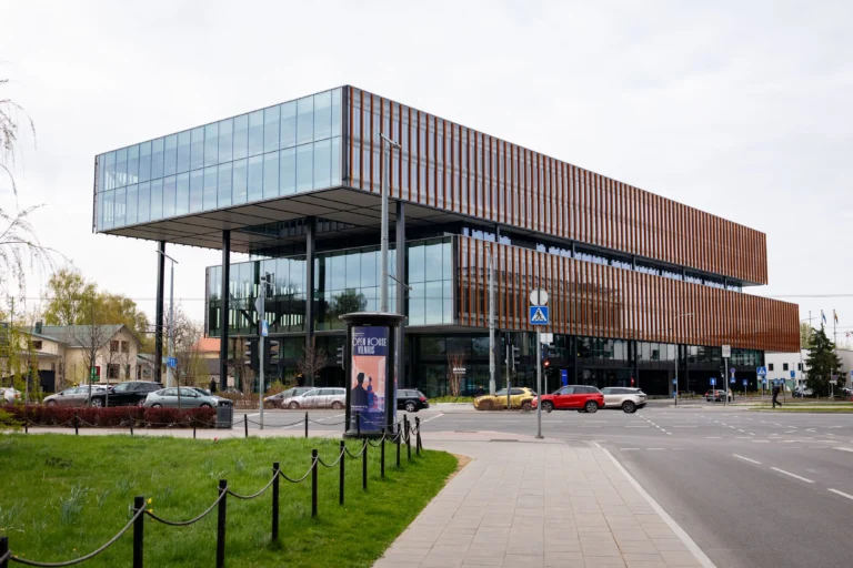 Moderna stiklota biroju ēka ar oranžām vertikālām līstēm un izvirzītu augšējo stāvu pie pilsētas ielas.