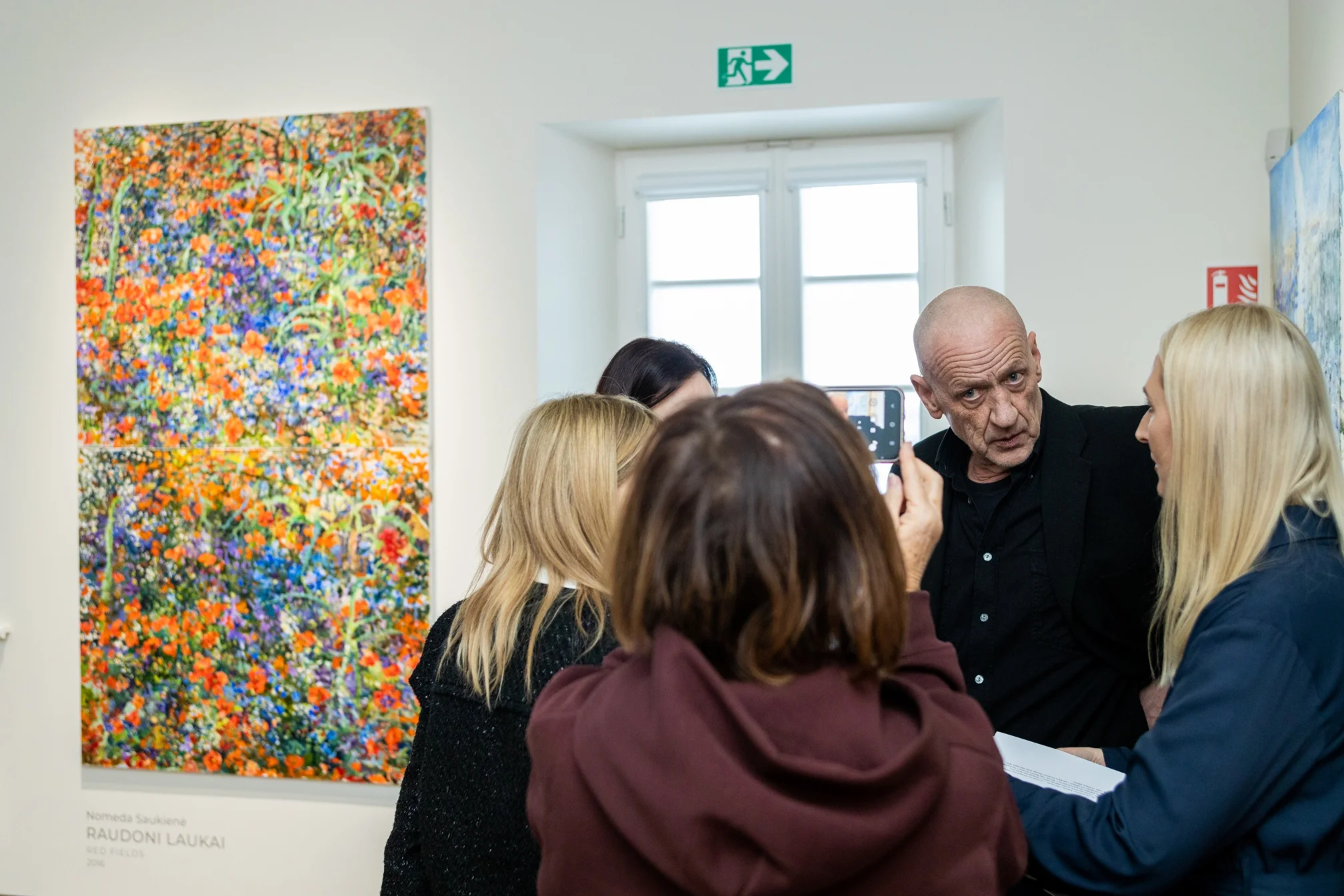 Vīrietis melnā apģērbā sarunājas ar sievietēm mākslas galerijā blakus košai, krāsainai ziedu gleznai.