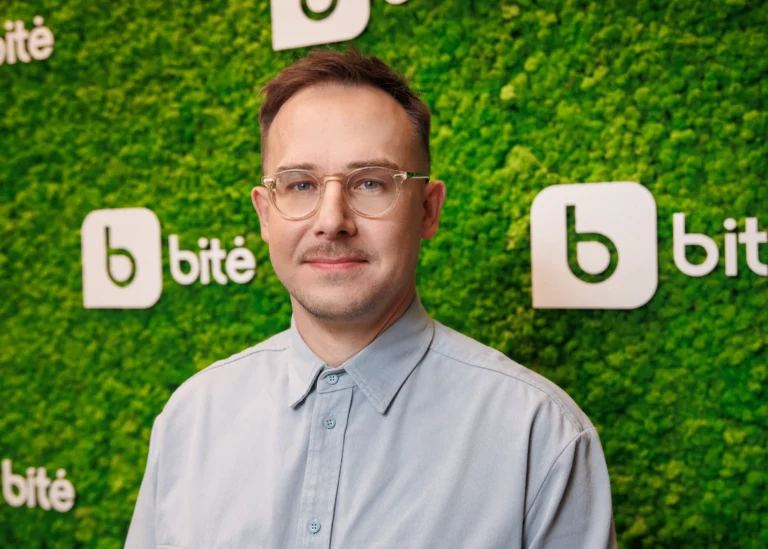 Vīrietis ar brillēm un gaiši zilu kreklu uz zaļas sūnu sienas fona ar Bite logotipu.