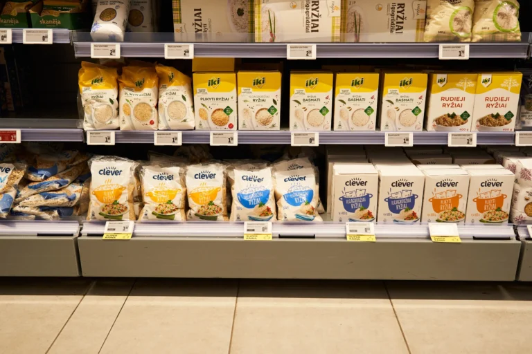Pārtikas veikala plaukti ar dažādu veidu rīsu iepakojumiem un redzamām cenu zīmēm apakšā.