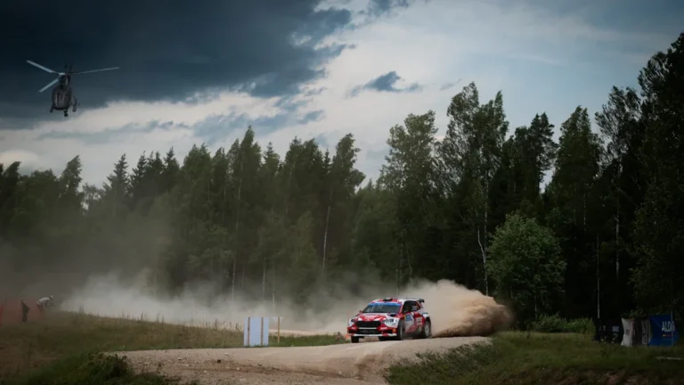 Rallija automašīna lielā ātrumā traucas pa zemes ceļu gar mežu, saceļot putekļu mākoņus.