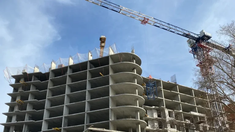 Daudzstāvu betona ēkas karkass būvniecības stadijā ar lielu celtni zilā debess fonā.