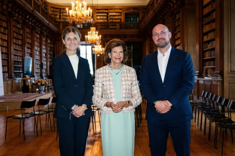 Karaliene Silvija stāv starp sievieti un vīrieti greznā, vēsturiskā bibliotēkā ar kristāla lustrām un grāmatu plauktiem.