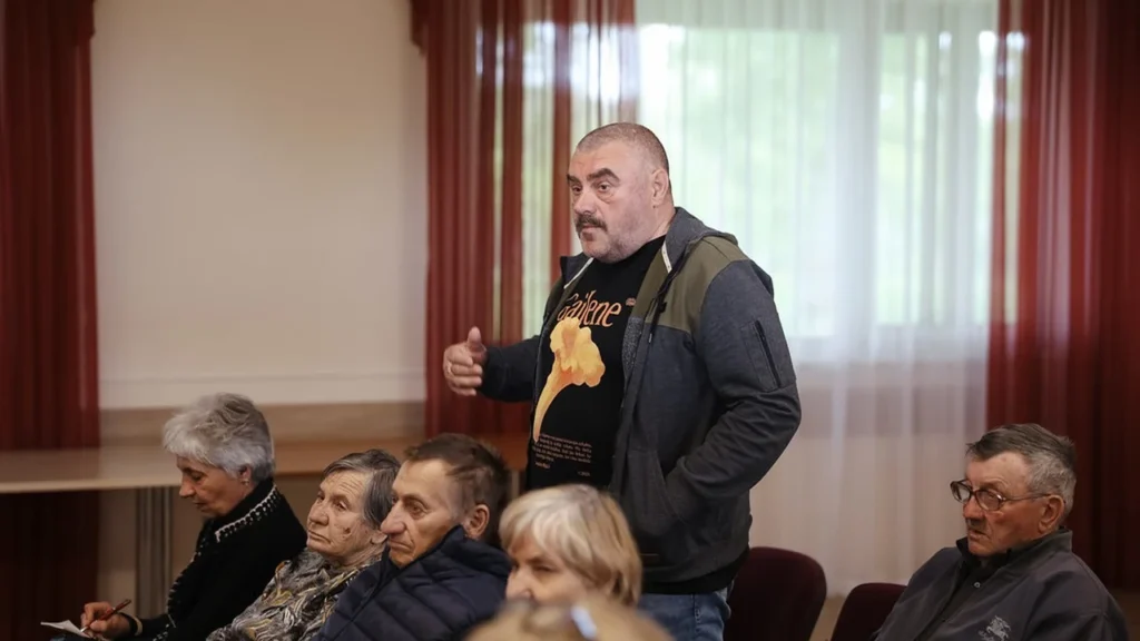 Vīrietis ar ūsām stāv un runā auditorijas priekšā, fonā sēž vairāki gados vecāki cilvēki.