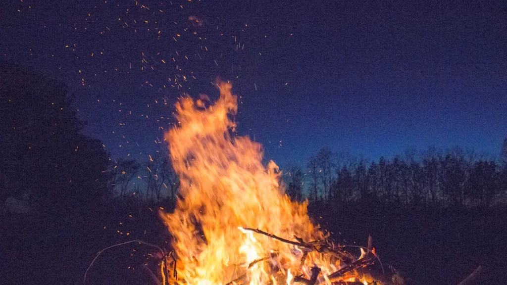Liels, spilgts Jāņu ugunskurs naksnīgā Latvijas lauku ainavā ar dzirkstelēm debesīs.