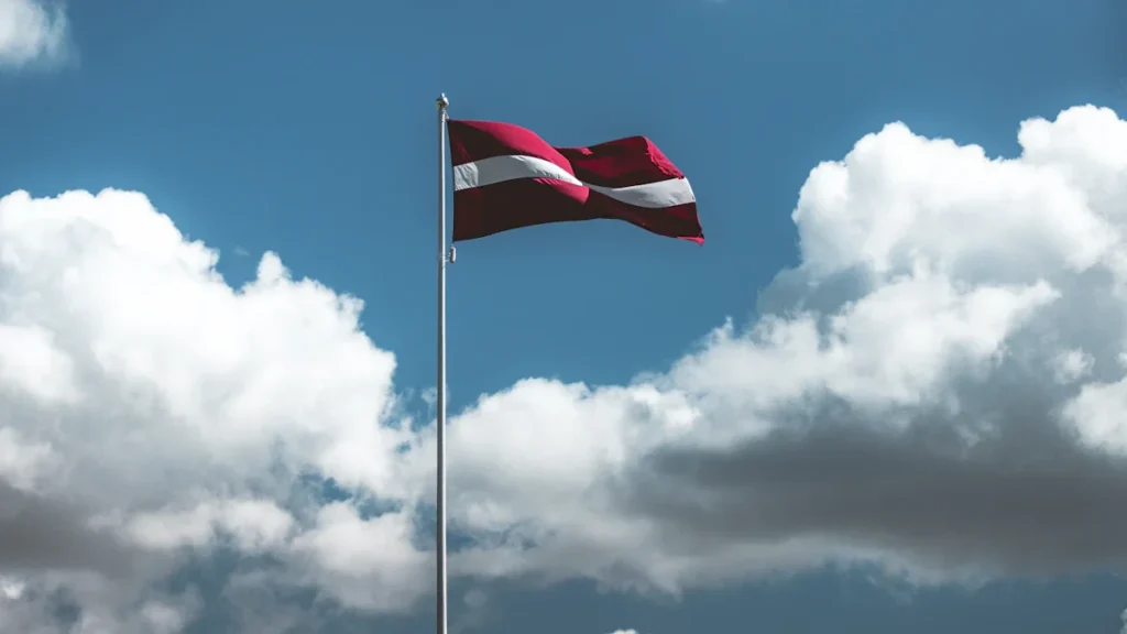 Latvijas valsts karogs plīvo zilas debesis ar mākoņiem fona.
