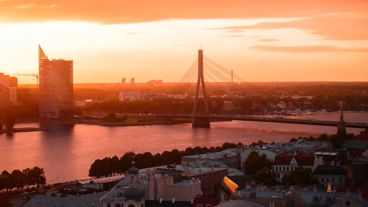 Rīgas pilsētas panorāma ar Vanšu tiltu zeltainā saulrietā, simbolizējot ekonomisko attīstību.