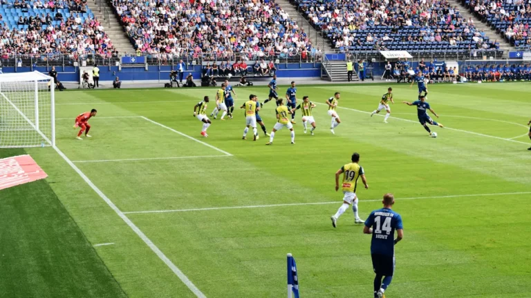 Futbola spēles norise stadionā, demonstrējot spēlētāju cīņu par bumbu laukumā.