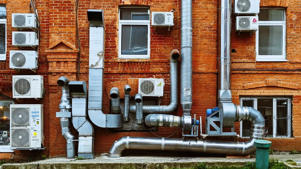Sarkanas ķieģeļu ēkas fasāde ar daudzām apkures un ventilācijas caurulēm.