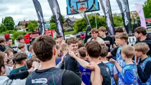 Jaunieši basketbola formās pulcējušies ap treneri brīvdabas laukumā ar Ghetto Basket reklāmas karogiem.