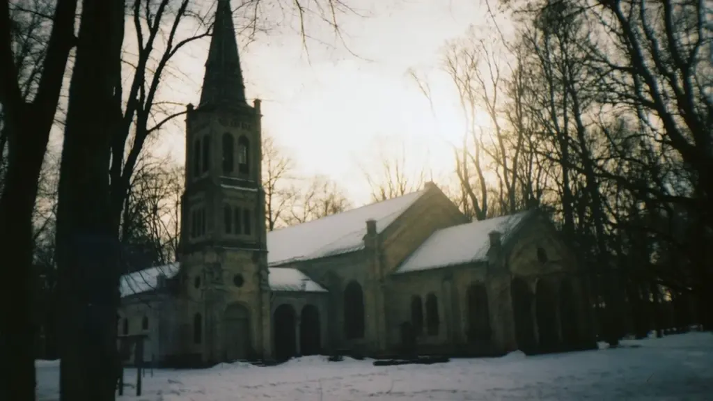 Vēsturiska baznīca kapsētas teritorijā ar smailu torni ziemas vakarā Latvijā.