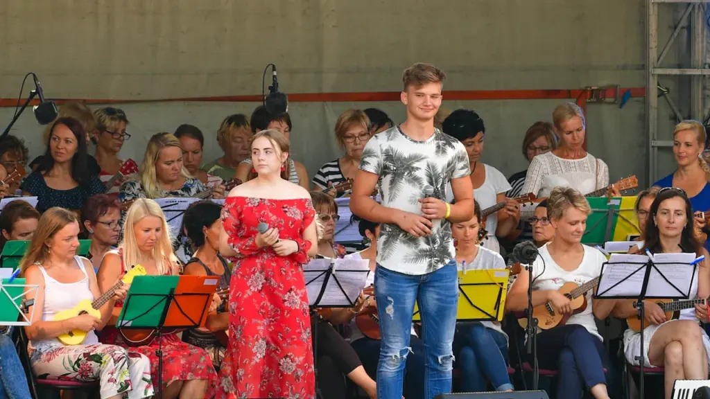 Jaunieši uzstājas uz skatuves, dziedot un spēlējot ukuleles lielā vasaras pasākumā brīvā dabā.
